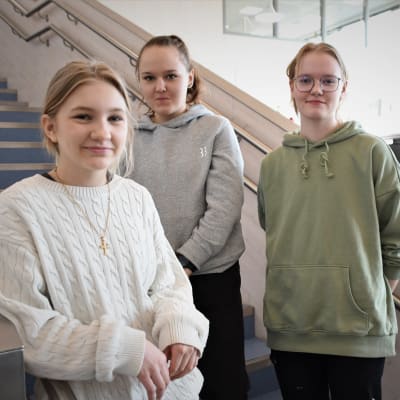Tre tonårsflickor står i en trappa inne i en skola.