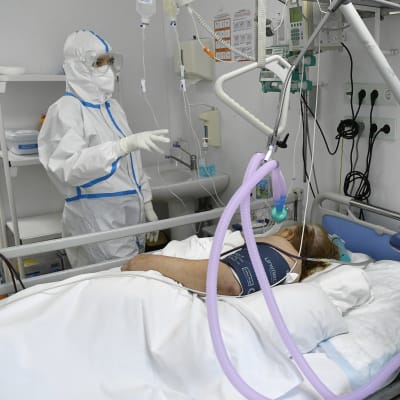En sjukskötare i skyddsutrustning står vid en sjukhussäng där en coronapatient ligger.