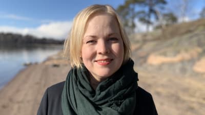 Närbild av en blond kvinna som står på en klippig strand i solskenet.