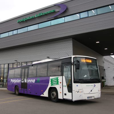 En lila och vit buss som står på en busstation. I bakgrunden en stor grå byggnad.
