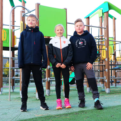 Tre grundskolelever står på rad framför en klätterställning.