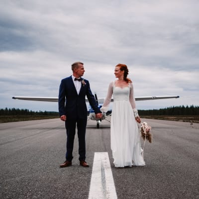 Hääpari seisoo lentokentän kiitotiellä toisiaan kädestä pidellen. Taustalla lentokone
