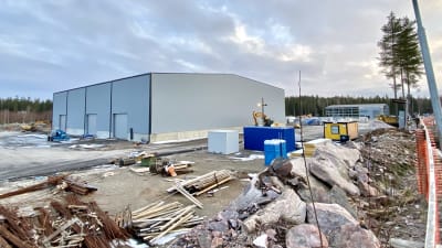 Industrihallar och byggmaterial på ett stort område. Stora stenblock, containrar, maskiner, stora ljusgrå hallar. Skog skymtar i bakgrunden.