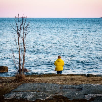 En kvinna i gul jacka står vid havet och tittar mot horisonten.