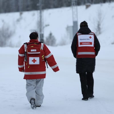 Kaksi henkilöä, joilla SPR:n liivit ja toisella myös reppu kävelee lumisessa maisemassa Vartiuksen raja-aseman lähellä.