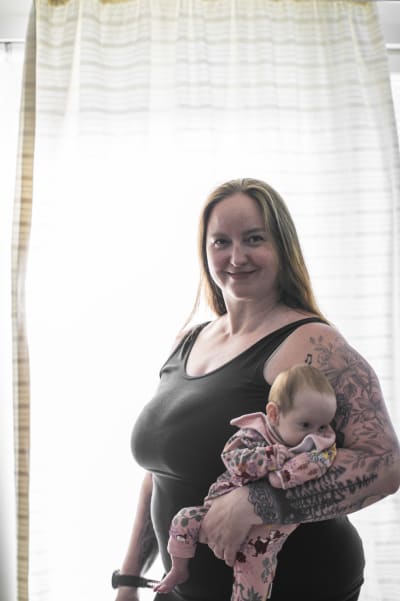 En kvinna står med en baby i famnen framför ett fönster med en gardin.