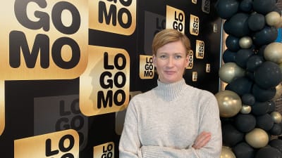 Päivi Rytsä står framför en affisch med Logomos emblem. 