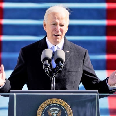 Biden puhuu puhujanpöntön takaa lavalla kongressitalon edustalla. Hän nostaa molempia käsiään.
