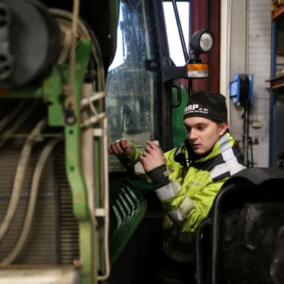 Nuori mies neonvärisessä työtakissa tarkistamassa traktorin öljyjä. 