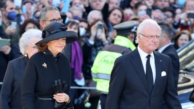 Drottning Silvia och kung Carl XVI Gustaf klädda i svart. Bakom dem syns en folkmassa där många håller upp mobiltelefoner.