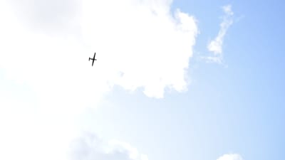 En drönade i luften, fotograferat från  marken. Drönaren är en svart siluett mot molnen.