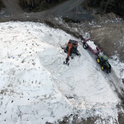 Ilmasta kuvattu kaivinkone lastaa lunta isosta lumikasasta traktorin lavalle.