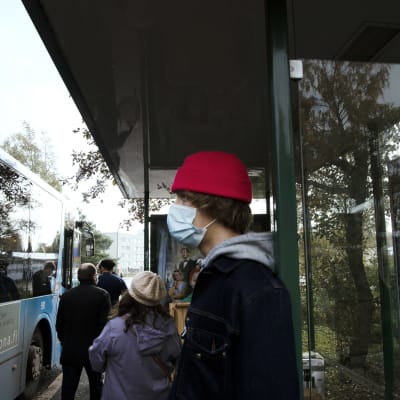 Kasvomaskiin pukeutunut henkilö seisoo bussipysäkillä.