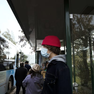 Kasvomaskiin pukeutunut henkilö seisoo bussipysäkillä.