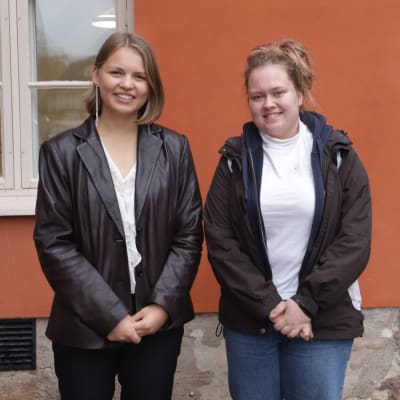 Två gymnasiestuderande vid Katedralskolan står framför en orange vägg. 