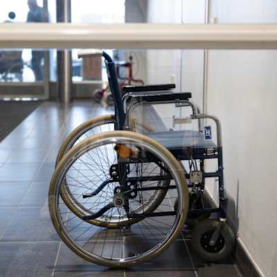 Tyhjä pyörätuoli rakennuksen sisääntuloaulassa, jossa valo tulvii sisään suurista lasiovista.