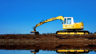 En grävmaskin gräver i torvjorden vid vattnet. Grävmaskinen speglas i vattenytan.