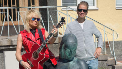 En man med långt blont hår och röd läderväst spelar på en gitarr. Berdvid honom en man som ler. De står vid en fontän.