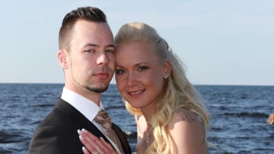 Nygift par står i bröllopsklädsel vid havet och ser in i kameran
