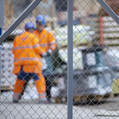 Två byggarbetare i orange overaller fotade genom ett nätstängsel