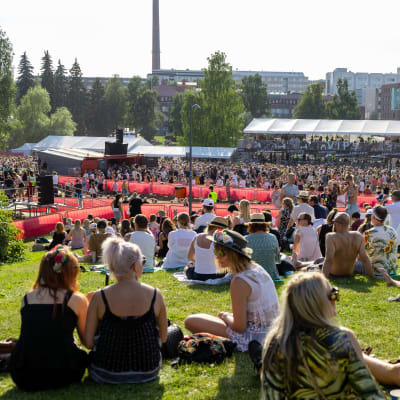 Tammerfest 2022 yleisöä Ratinanniemessä. Ihmiset istuvat nurmella etualalla. Taustalla VIP -alue ja paljon ihmisiä katsomassa konserttia.