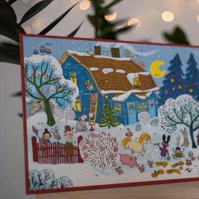 Kuvassa on adventtikalenteri pöydällä. Kalenterissa on piirroskuva talvisesta maisemasta, jossa näkyy muun muassa sininen talo, lunta, puita ja eläimiä.