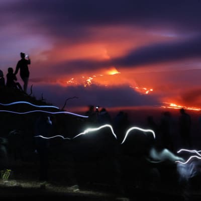 ihmiset katsovat tulivuorenpurkausta yöllä, kännyköiden lampuista valojuovia