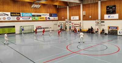 Kuvassa harmaalattiainen sisäpelihalli, jossa kesken pelitilanteen valko- ja punapaitaisia futsal-pelaajia.