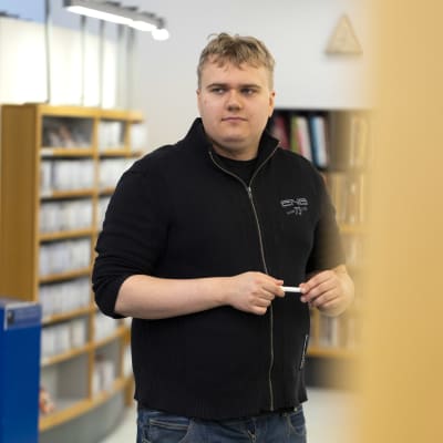 Opiskelja Jussi Hartikainen seisoo kirjastossa. Taustalla näkyy sumeana kirjahyllyjä.