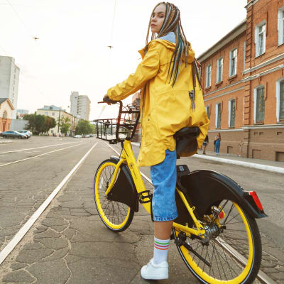 En kvinna i en gul regnrock sitter på en cykel. I bakgrunden byggnader vid en asfalterad väg.