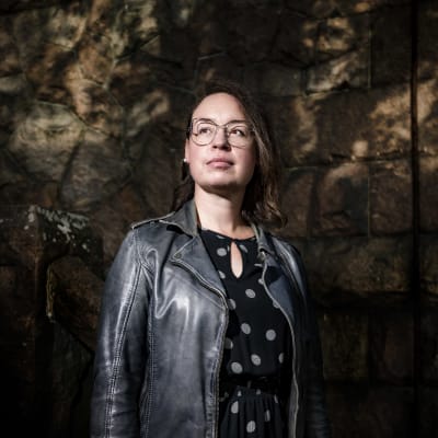 Kvinna står och tittar åt sidan, mot en mörk bakgrund.