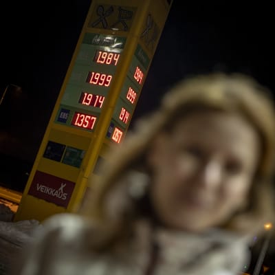 Minna Jaala seisoo huoltoaseman pihalla. Taustalla polttoaineiden hinnat. 