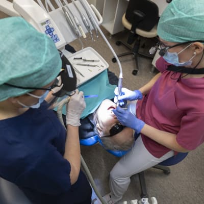 Två personer sköter om tänderna på en äldre man i tandläkarstol.