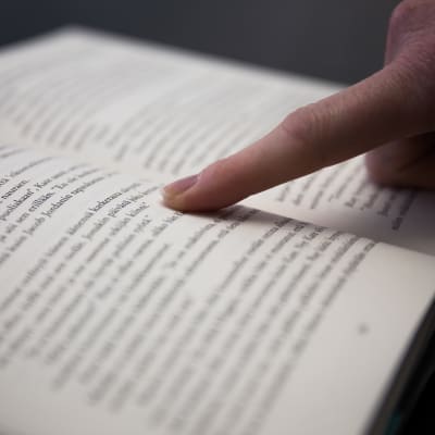 avoinainen kirjan aukeama tummalla taustalla, lukija seuraa tekstin rivejä sormellaan