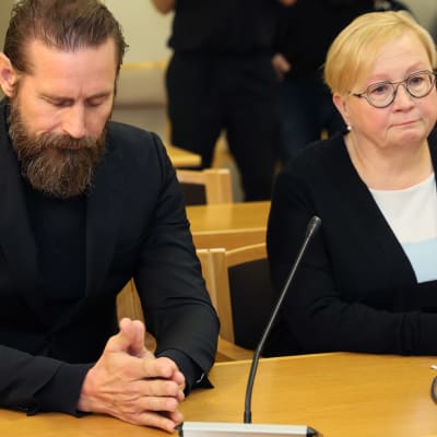 Mustiin pukeutunut, parraskas laulaja Lauri Tähkä ja asianajajansa Riitta Leppiniemi lähikuvassa istumassa oikeudessa päydän takana. Tähkän katse luotu alas, kädet yhdessä pöydällä.