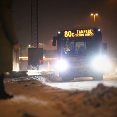 Ihmisiä odottamassa bussia lumisateessa.