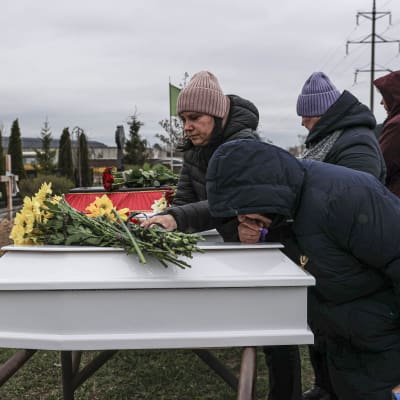 Naiset surevat lasten hauta-arkkujen vierellä, yksi naisista on kumartunut arkun yllä, toinen asettelee kukkia arkun päälle.