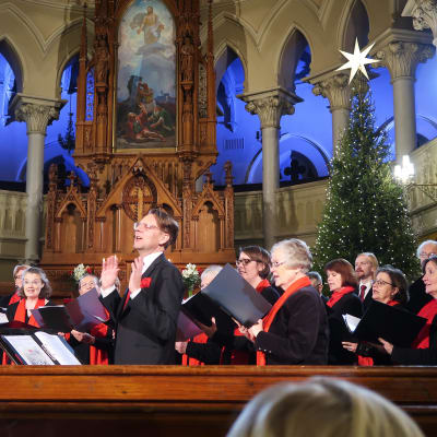 Kör sjunger klädd i svart och rött framme vid träaltaret i den i övrigt vita Johanneskyrkan. I bakgrunden julgran med stjärna i topp.