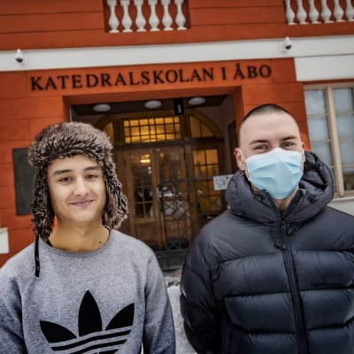 Till vänster en ung man med grå collegetröja och pälsmössa och till höger en ung man med mörkblå vinterrock och munskydd.