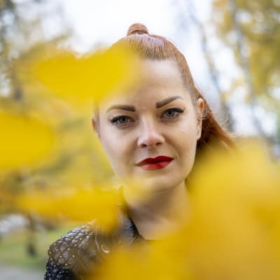 Nainen katsoo suoraan kameraan syksyisten lehtien takaa
