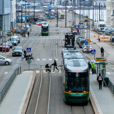 Laajassa kaupunkimaisemassa näkyy raitiovaunuja, lastenvaunujen kanssa suojatietä ylittäviä henkilöitä, autoja ja pyöräilijöitä liikkeessä.