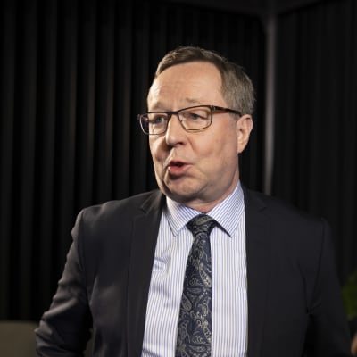 Silmälasipäinen mies puhuu ohi kameran mustaa taustaa vasten seisten.