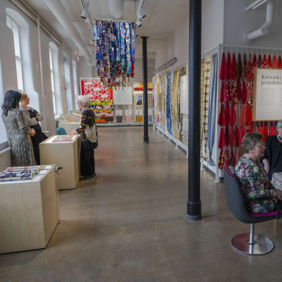 Museotila. Etualalla on pöytä, jonka ääressä istuu kolme vanhempaa naista. Vasemmalla isojen ikkunoiden edessä seisoo myös muutamia naisia. Seinillä ja katosta roikkuu kirkkaanvärisiä kankaanpaloja.  