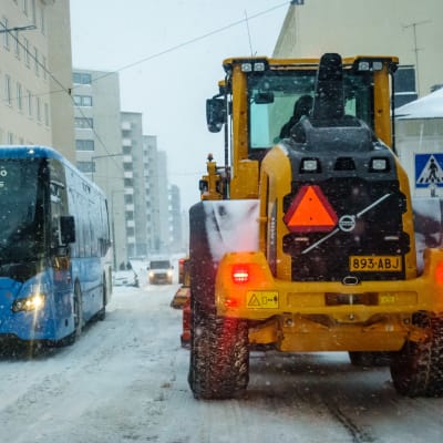 En snöplog och en buss möts på gatan i snöoväder. 