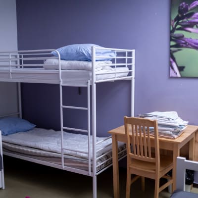 Den här bilden visar ett familjerum på ett skyddshem i Helsingfors. Det syns en våningssäng och ett skrivbord och en stol på bilden. Väggen är violett och det hänger en tavla ovanför bordet.