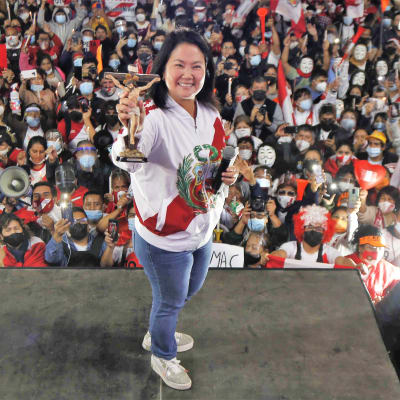 Keiko Fujimori seisoo hymyillen puhujanlavalla esitellen pientä patsasta, joka esittää Jeeusta ristillä. Toisessa kädessään hänellä on mikrofoni. Hänellä on yllään valkoinen pusakka ja siniset farkut. Taustalla näkyy yleisömerta.