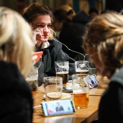Ett gäng kvinnor sitter i en bar i Paris med halvdruckna öl på bordet framför sig. De håller upp sina mobiler och tittar och lyssnar på Macrons tal om en ny nedstängning.