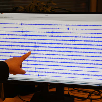 Seismologi osoittaa sormella sinisiä mittauskäyriä tietokoneen näytöllä. Taustalla kellertävä valo toimistohuoneessa.