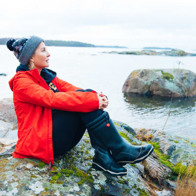Kvinna i röd jacka sitter på en klippa och ser ut över havet.