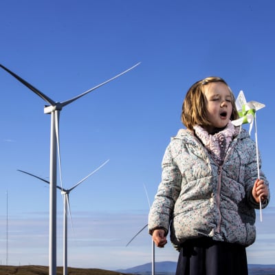 Kolme suurta tuulimyllyä seisoo maisemassa sinistä taivasta vasten. Etualalla pikkutyttö puhaltaa kädessään olevaan tuulihyrrään.